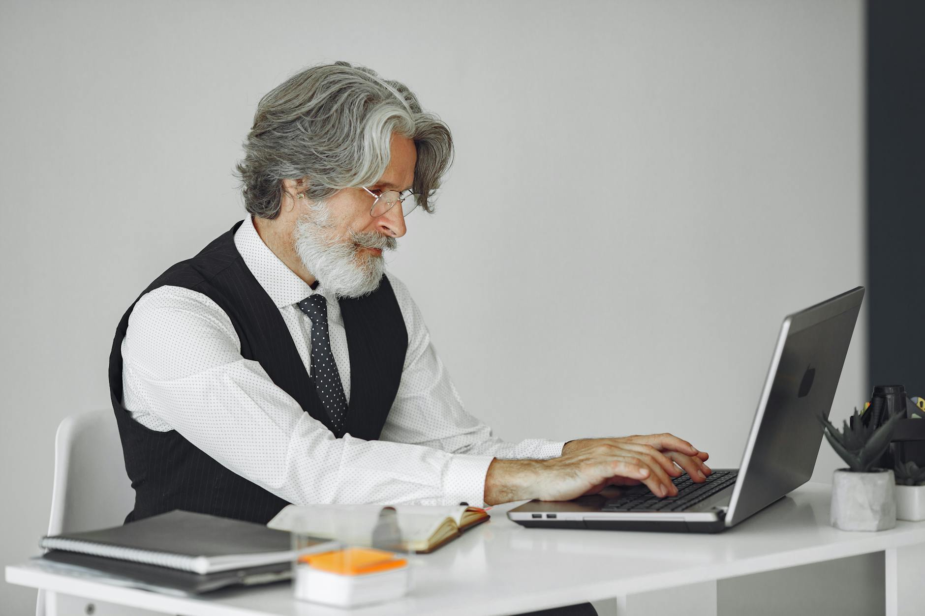 Elegant senior man in office attire typing on a laptop at a desk.