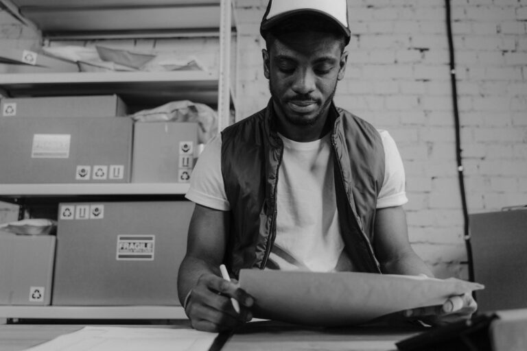 Warehouse worker in a cap packaging a parcel with boxes in the background.