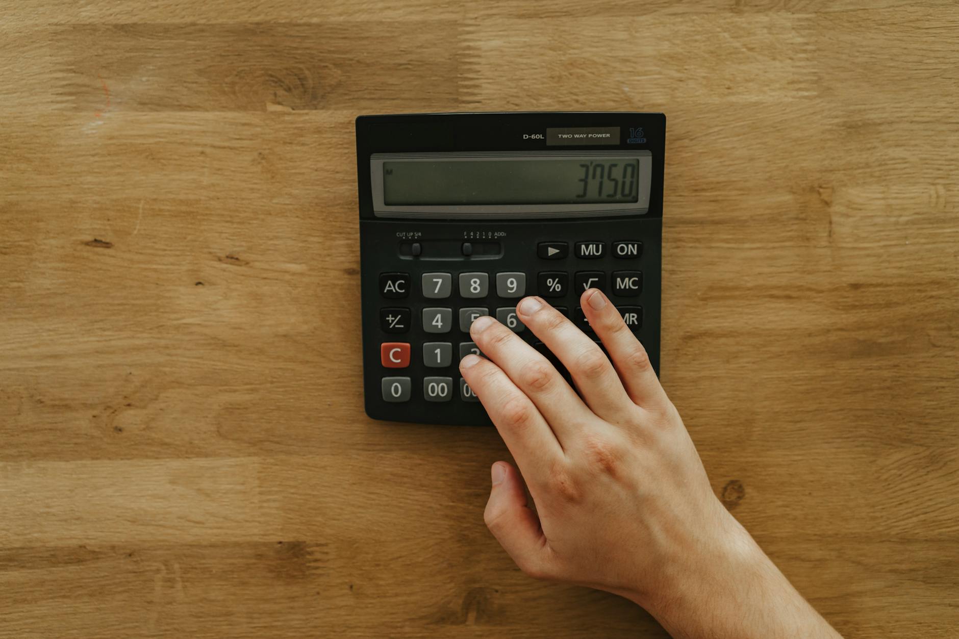 A person's hand pressing keys on a calculator displaying 3750 on a wooden table, top view.
