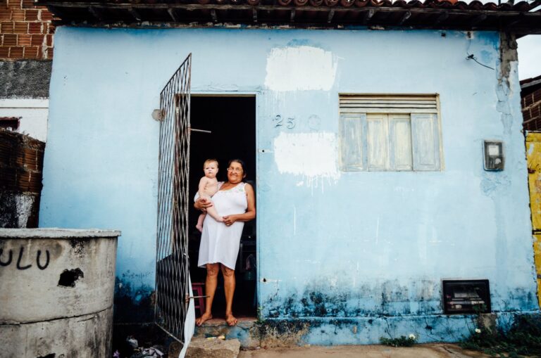 A cheerful woman holding a baby at the entrance of a blue rustic house in Natal, Brazil.