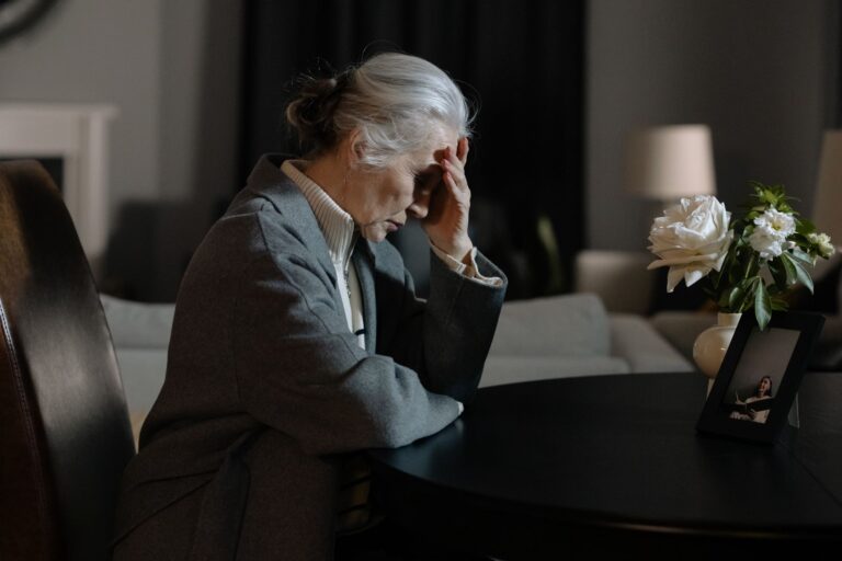 An elderly woman sitting indoors, hand on face, next to a bouquet and framed photo, evokes a reflective mood.