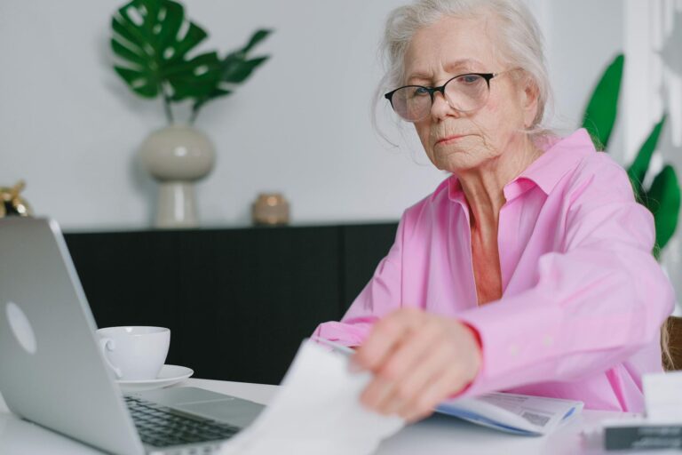 Elderly woman in pink shirt working on a laptop at home, focused and engaging with documents.