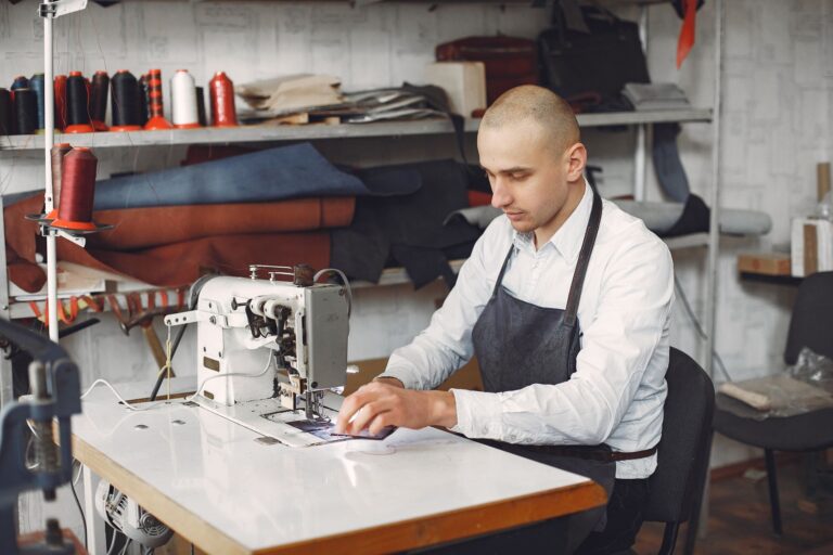 Side view of concentrated adult male artisan in formal clothes and apron using sewing machine while working in studio