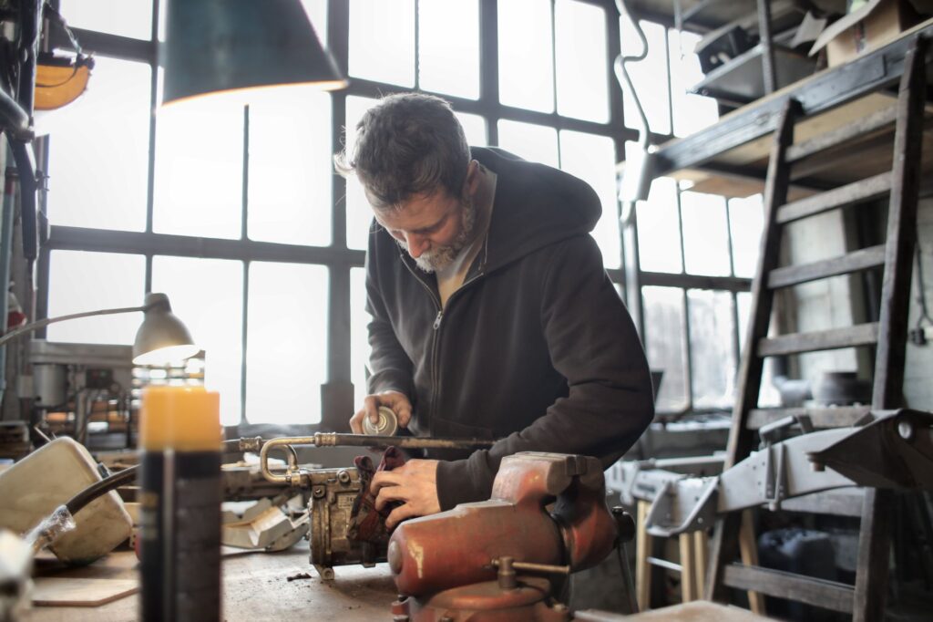 Male craftsman in casual outfit fixing old rusty detail in workshop and using aerosol paint while standing at workbench and working