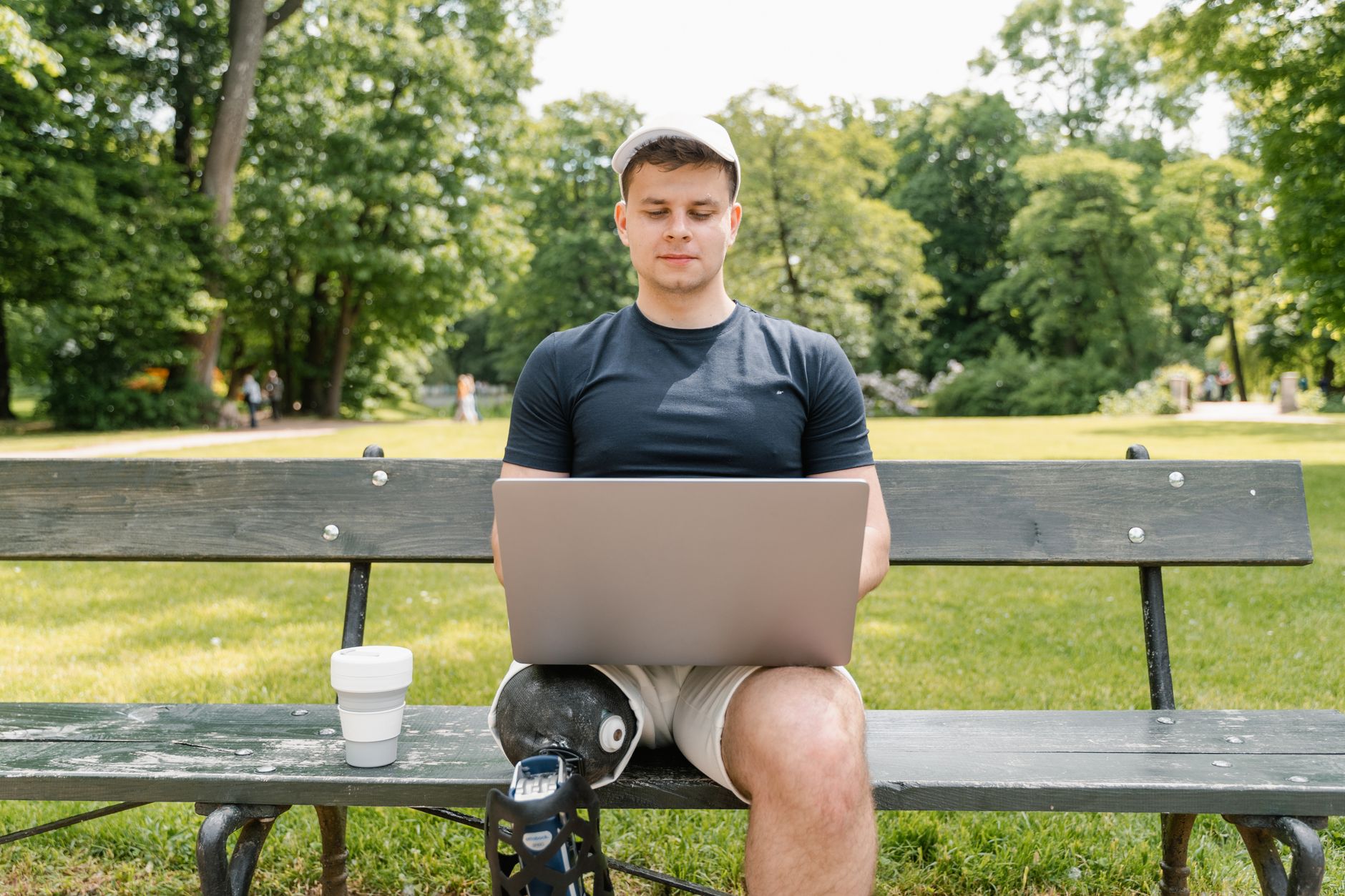 Young man with prosthetic leg works on a laptop while sitting on a park bench, featuring a serene outdoor setting.