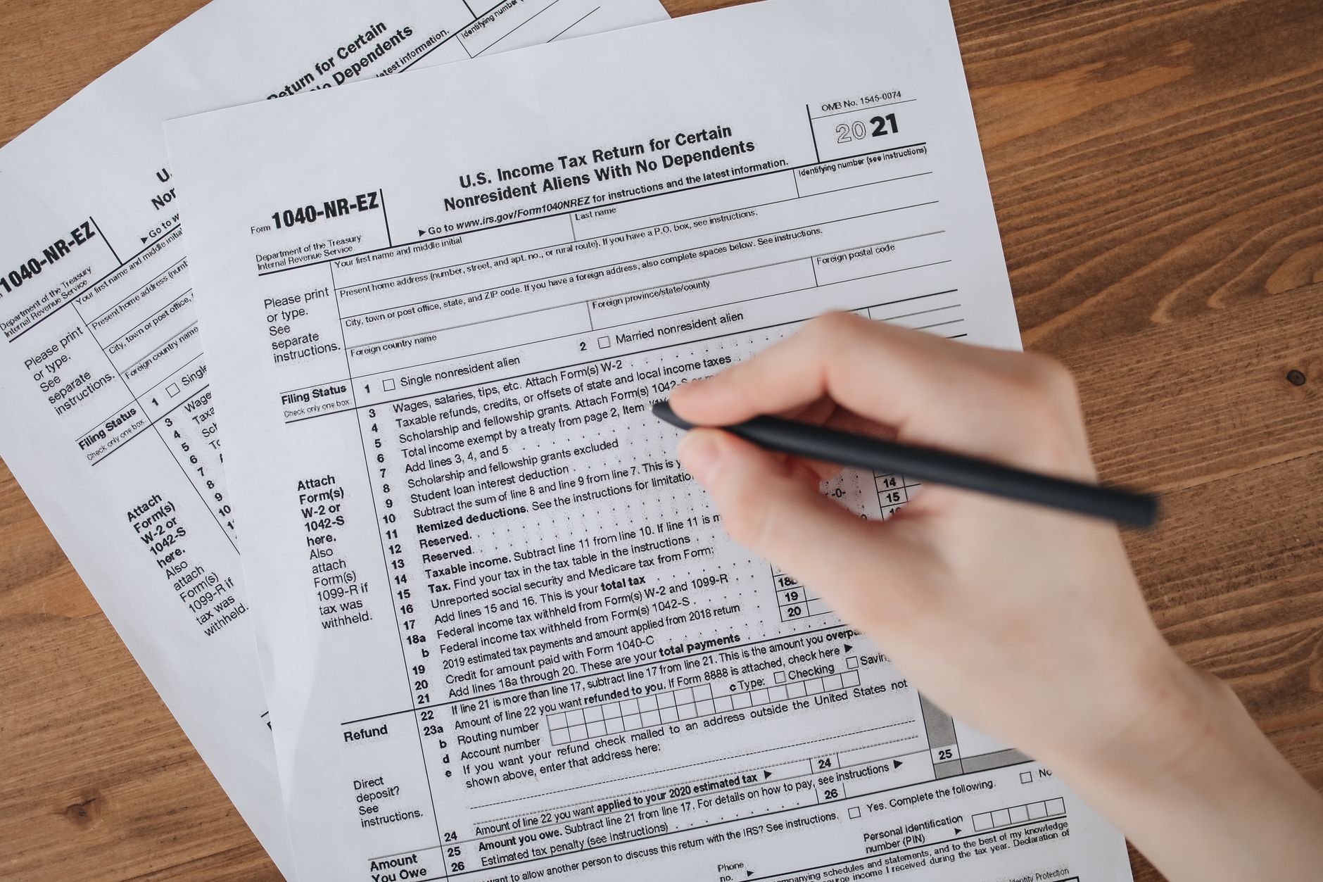 Person filling out U.S. tax form 1040-NR-EZ with a pen on a wooden table.