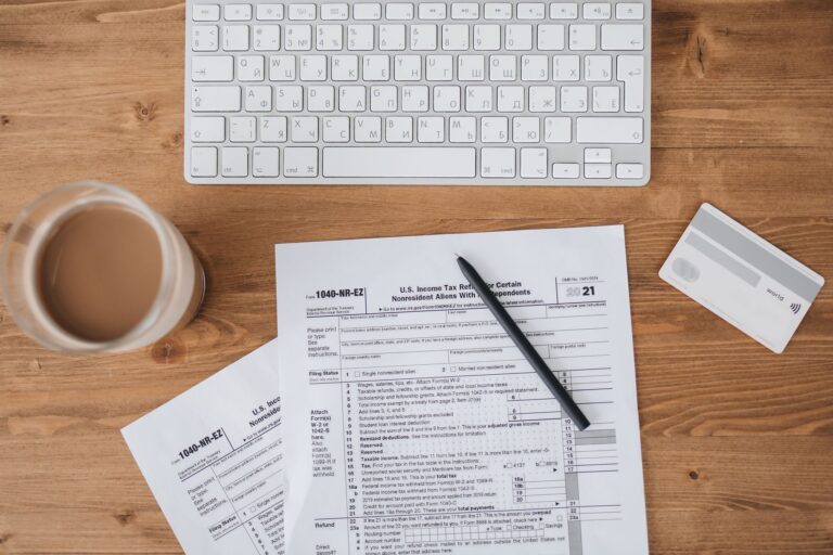 Top view of tax forms, keyboard, pen, and coffee on a wooden desk.