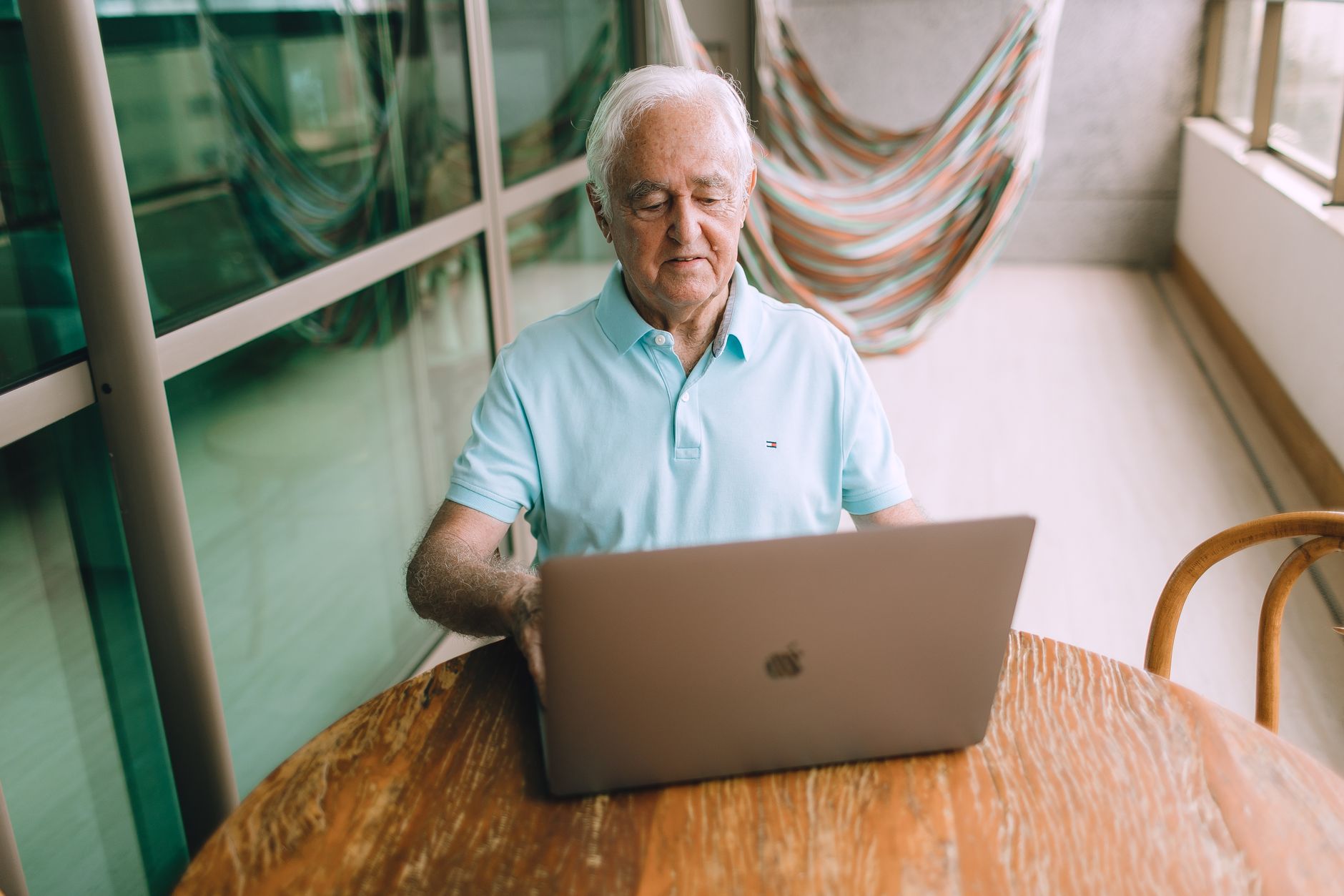 Elderly man seated at a table on a balcony, surfing the internet with a laptop.