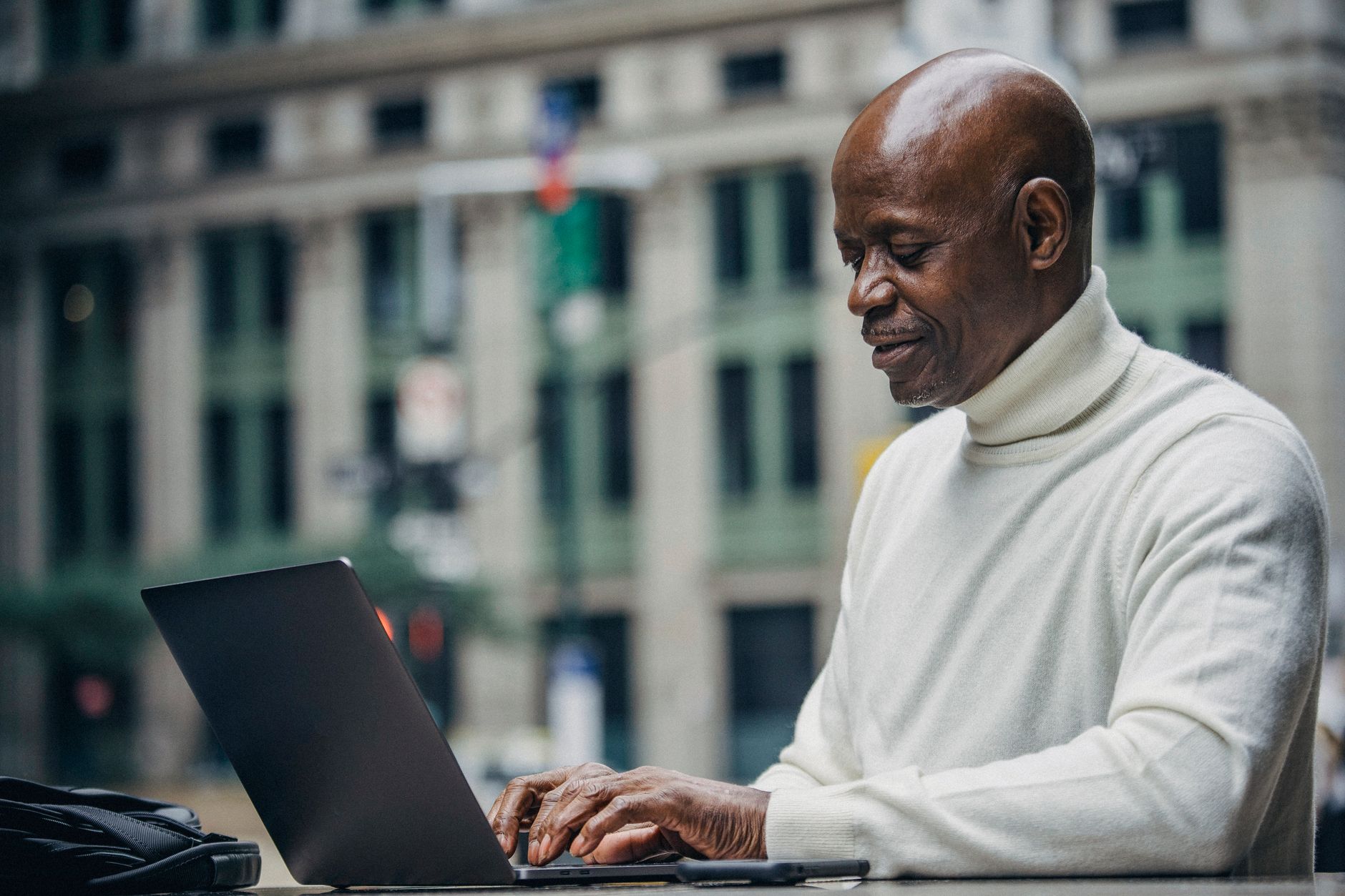Side view of self employed concentrated mature bald black man in stylish turtleneck sitting in street cafe and typing on netbook during work