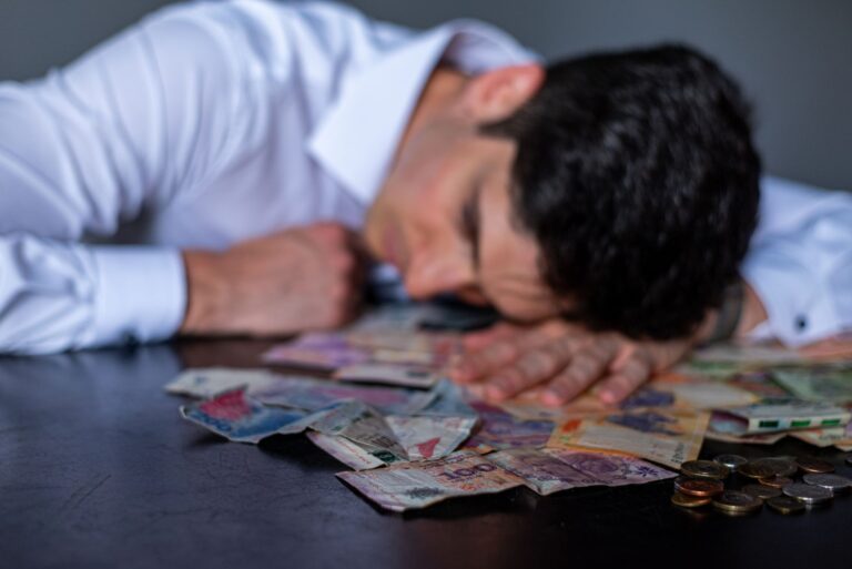 A man in a white shirt rests his head on a table covered with Argentine pesos and coins.