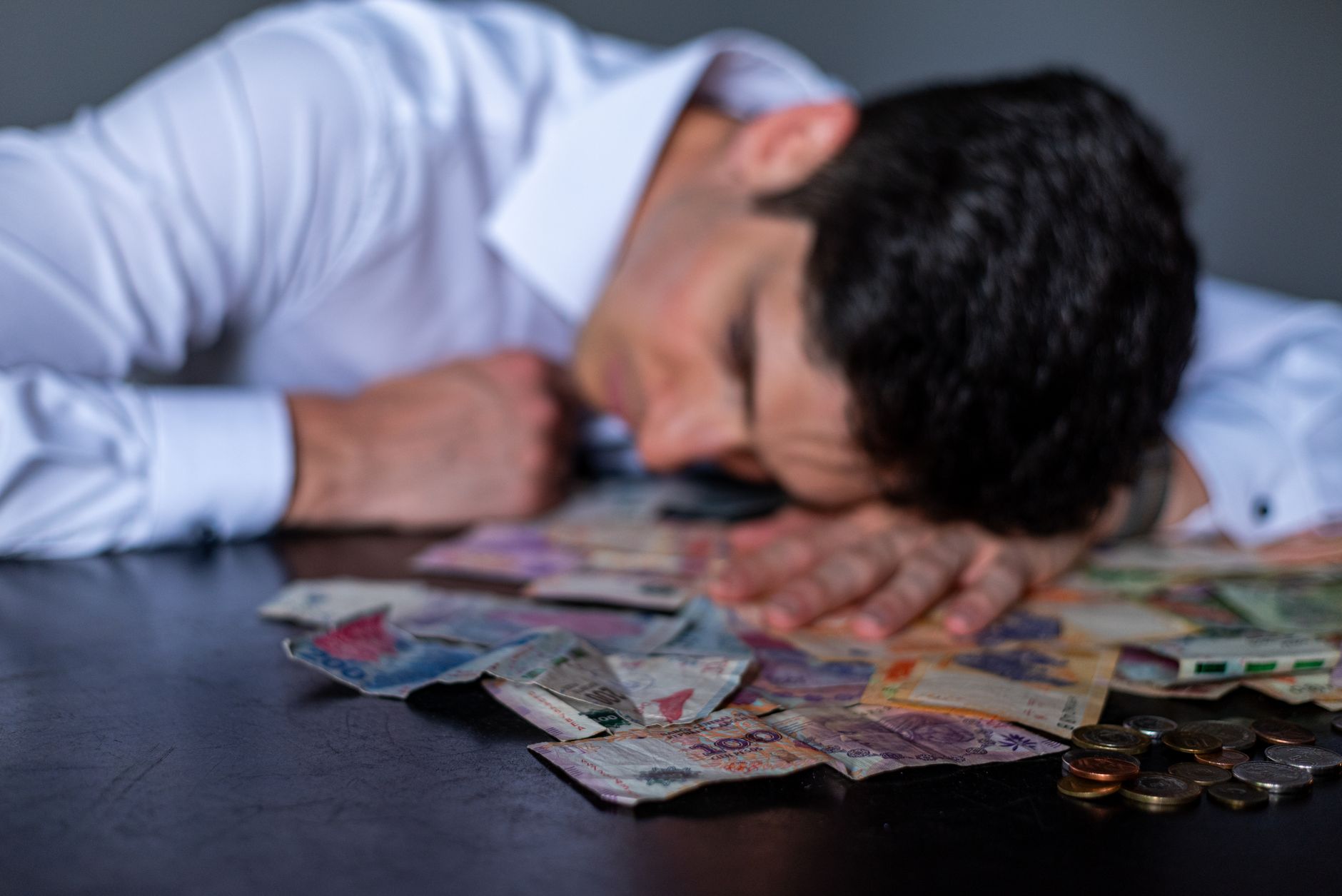 A man in a white shirt rests his head on a table covered with Argentine pesos and coins.