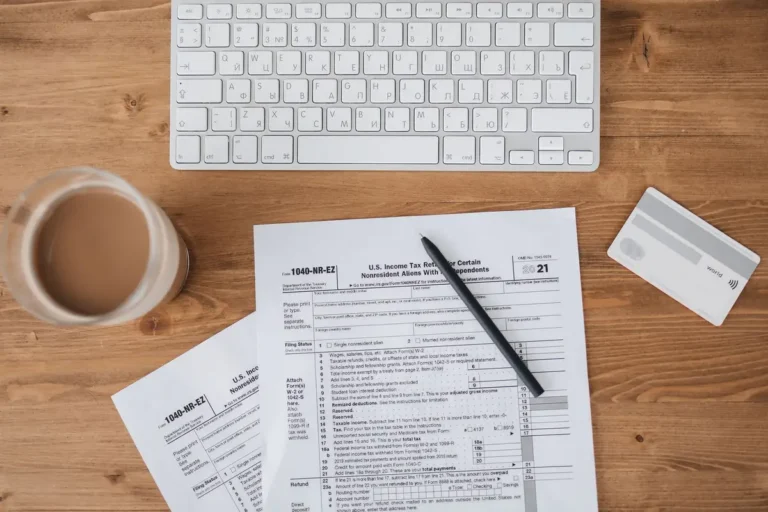 Top view of tax forms, keyboard, pen, and coffee on a wooden desk.