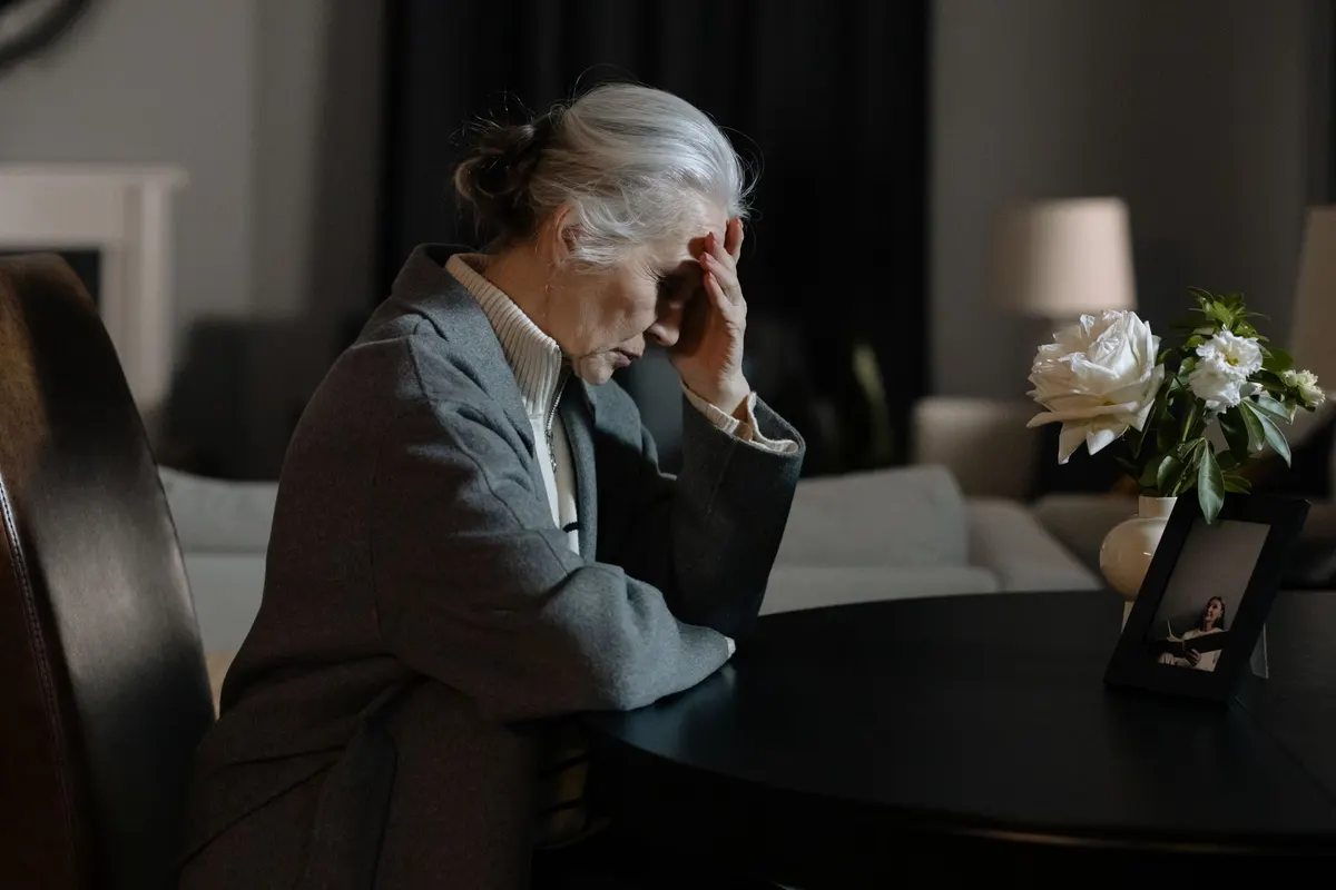 An elderly woman sitting indoors, hand on face, next to a bouquet and framed photo, evokes a reflective mood.