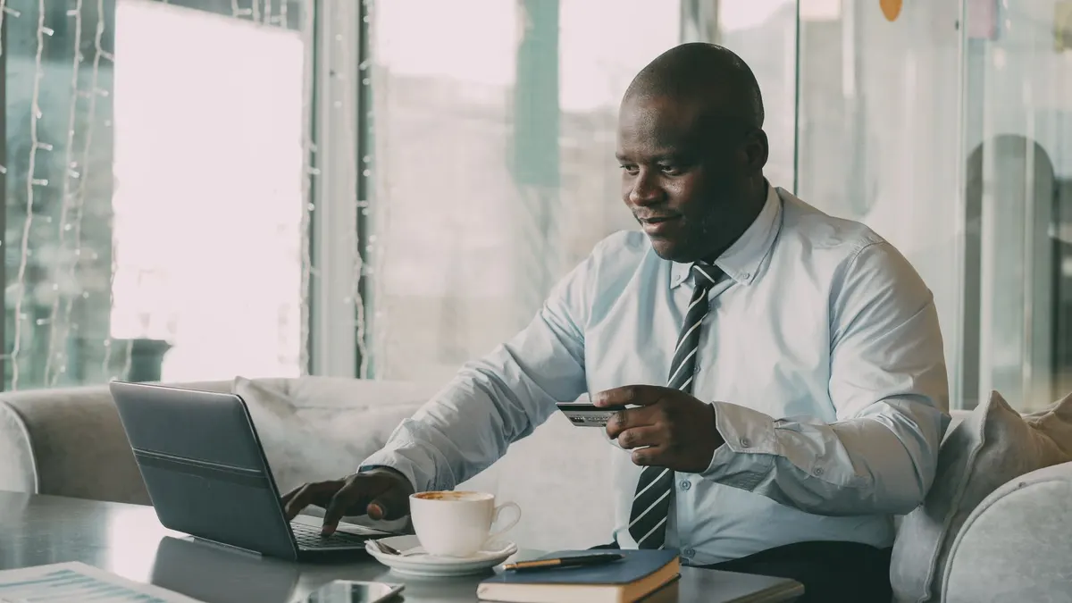 Black businessman using a laptop and credit card in a modern cafe setting.