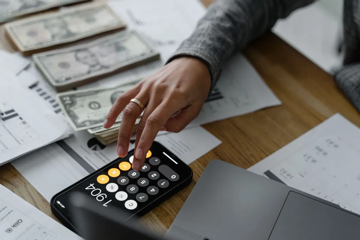 Close-up of a person using a smartphone calculator amid money and financial documents on a wooden table.
